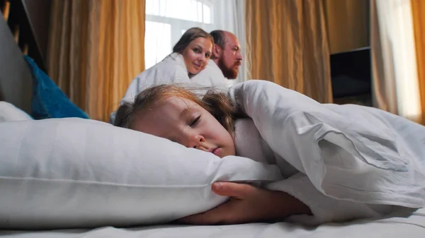 A family in the hotel room - a little girl sleeping in bed - smiling mom and dad looking at her — Stock Photo, Image