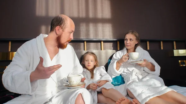 Rodina v bílých županech sedí v posteli a ráno pije čaj. — Stock fotografie