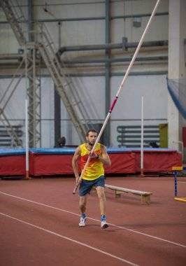Kapalı alanda sırıkla atlayan sarı gömlekli bir adam pistte sırıkla dikiliyor.