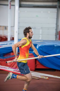 Pole kapalı alanda atlıyor - atletik bir adam spor stadyumunda bir direkle koşuyor.