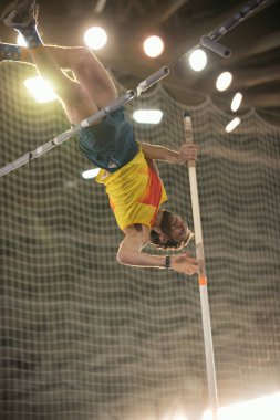 Spor stadyumunda sırıkla atlama. Sarı tişörtlü bir adam barın üzerinden atlıyor.