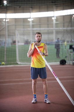 Spor stadyumunda sırıkla atlama. Sarı gömlekli bir adam kameraya poz veriyor.