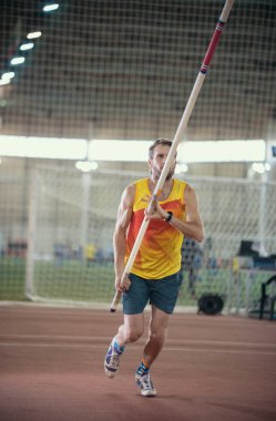 Spor stadyumunda sırıkla atlama. Sarı tişörtlü atletik bir adam pistte koşuyor.