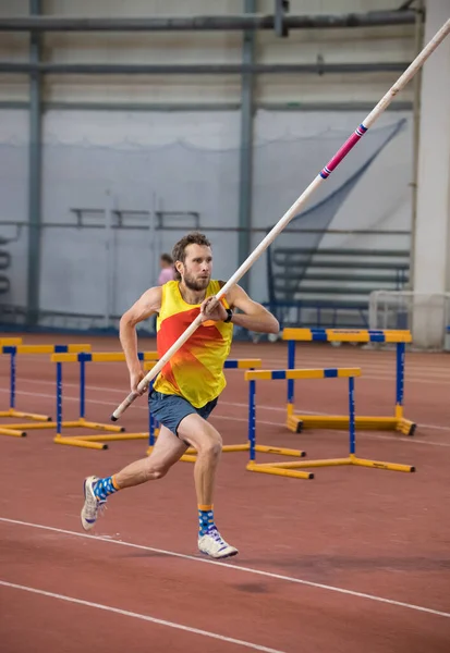 Kapalı alanda atlayan sarı tişörtlü atletik bir adam stadyumda bir direkle koşuyor. — Stok fotoğraf