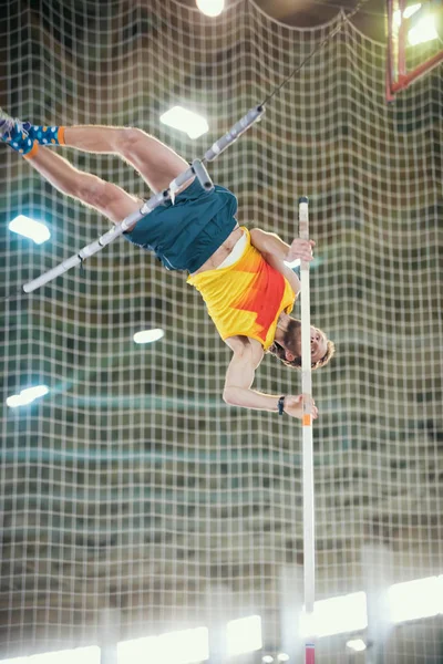 Stabhochsprung drinnen - ein sportlicher Mann springt über die Stange - gestützt auf die Stange — Stockfoto