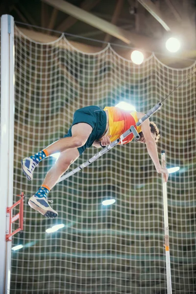 Stabhochspringen drinnen - ein sportlicher Mann springt über die Stange - an die Stange gelehnt - helles Licht im Hintergrund — Stockfoto