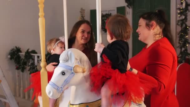 Concepto de Navidad - Dos bebés sentados en caballos de carrusel y jugando con ellos - sus madres hablando entre sí — Vídeo de stock