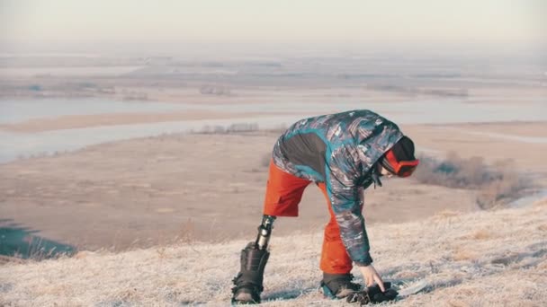 Сноуборд - человек с протезной ногой стоит на доске и надевает перчатки — стоковое видео