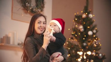 Noel konsepti - Küçük bebeğini Noel stüdyosunda tutan gülümseyen genç bir kadın - oyuncak geyik tutan bir bebek