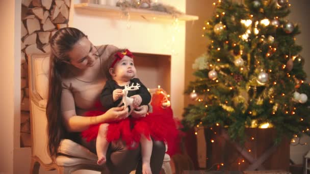 Concetto natalizio - Una giovane donna sorridente seduta sulla sedia con la sua figlioletta nello studio di Natale - che gioca con un cervo giocattolo — Video Stock