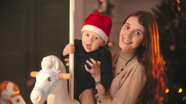 クリスマスのコンセプト-赤ちゃんがカメラを見ている若い母親-赤ちゃんがカルーセルに座って — ストック動画