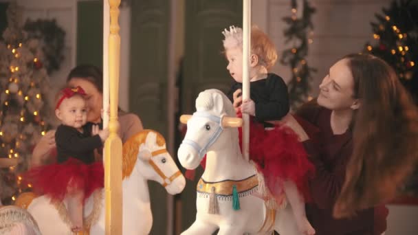 Estudio decorado de Navidad - Dos madres sonrientes divirtiéndose con sus bebés sentados en el carrusel — Vídeos de Stock