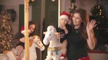 Noel konsepti - iki bebek atlıkarıncada oturup onlarla oynuyor - anneleri onları tutuyor ve kameraya el sallıyor