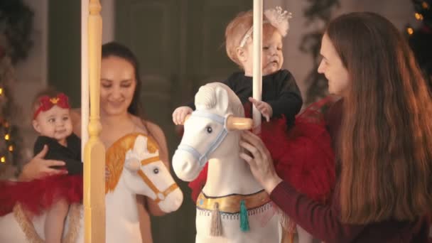 Kerst ingerichte studio - Twee lachende gelukkige moeders hebben plezier met hun baby 's zitten op carrousel — Stockvideo
