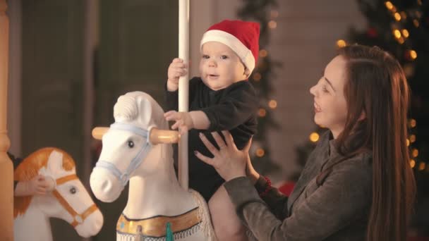 Різдвяна прикрашена студія - усміхнена молода мати стоїть поруч зі своєю дитиною на каруселі — стокове відео