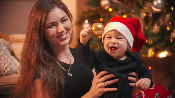 Concepto de Navidad - Una mujer sonriente sosteniendo a su pequeño hijo - el bebé jugando con la bola de navidad dorada — Foto de Stock