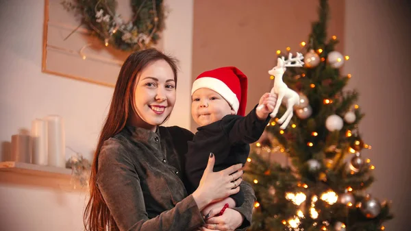 Concepto de Navidad - Una joven sonriente sosteniendo a su pequeño bebé en el estudio de Navidad - un bebé sosteniendo ciervos de juguete — Foto de Stock