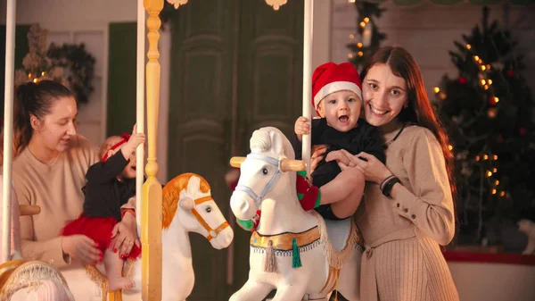 Divertirse con la decoración de Navidad - dos madres sonrientes de pie cerca del carrusel con sus bebés — Foto de Stock