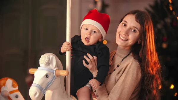 クリスマスのコンセプト-赤ちゃんがカメラを見ている若い母親-赤ちゃんがカルーセルに座って — ストック写真