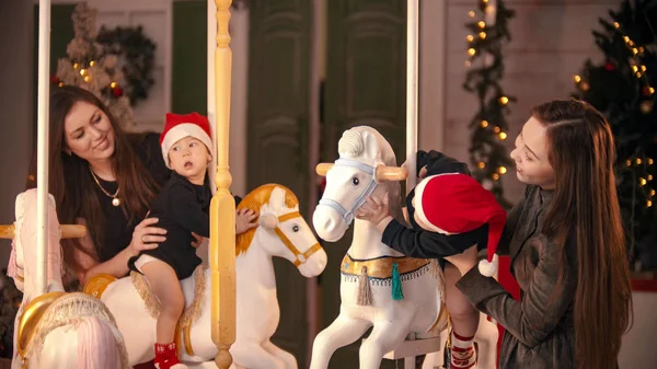 クリスマスのコンセプト- 2人の赤ちゃんがカルーセル馬に屋内に座って一緒に遊んでいます-母親がそれらを保持 — ストック写真