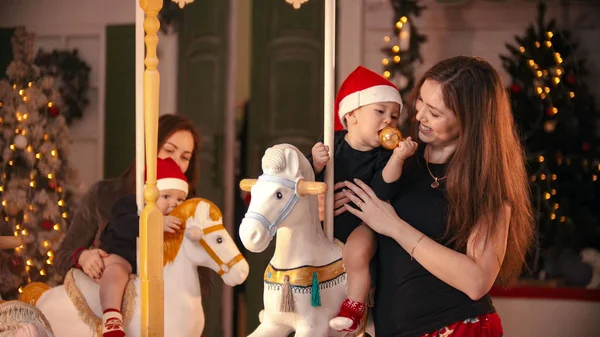 Christmas concept - Two babies sitting on carousel horses indoors and playing with them - a baby chewing a christmas ball — ストック写真