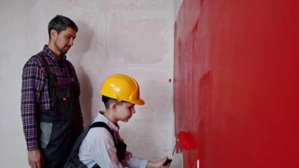 Ein kleiner Junge im Helm bemalt Wände mit roter Farbe - sein Vater beobachtet ihn — Stockvideo