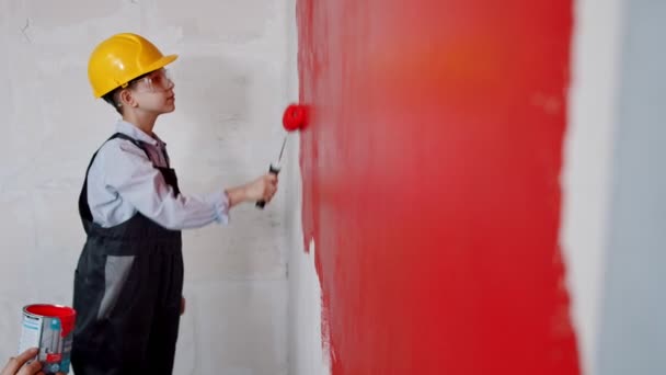 Renovación del apartamento - un niño pequeño con casco y gafas pintando la pared en rojo — Vídeos de Stock