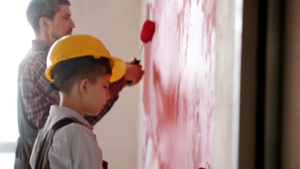 Een kleine jongen en zijn glimlachende vader schilderen muren in rode kleur - een jongen met een helm op — Stockvideo