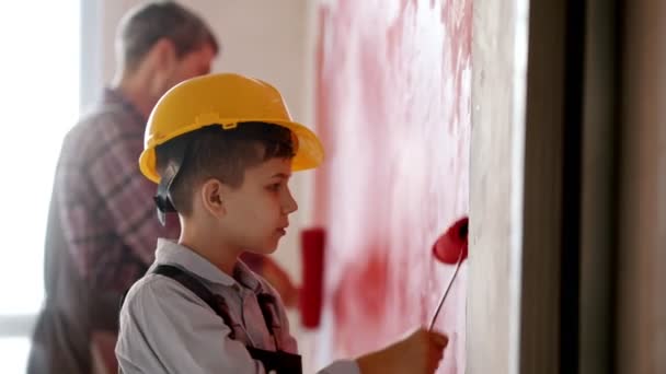 Küçük bir çocuk ve gülümseyen babası yeni dairenin duvarlarını kırmızıya boyuyor. Kasklı bir çocuk. — Stok video
