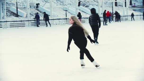 A young blonde woman professional figure skater skating on the outdoors ice rink around people — ストック動画