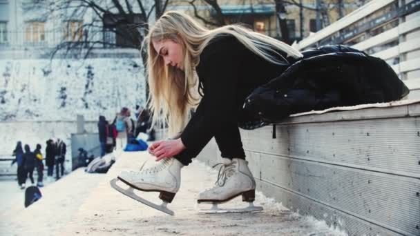 Sarı saçlı genç bir kadın buz pistine çıkmadan önce buz patenlerini bağlıyor. — Stok video