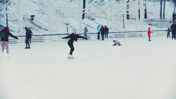 A young blonde woman skating on ice rink around other people outdoors — 스톡 사진