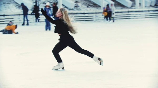 Eine junge blonde attraktive Eiskunstläuferin, die auf der Eisbahn im Freien um Menschen herum Schlittschuh läuft — Stockfoto