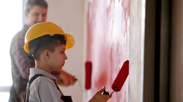 Een kleine jongen en zijn glimlachende vader schilderen muren in rode kleur - een jongen met een helm op — Stockfoto