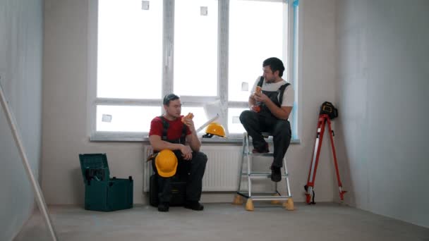 Apartman javítása a draft apartman - férfi munkavállalók, akik egy kis szünetet, és eszik sajtos tekercs — Stock videók