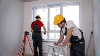 Apartment repair in the room - two men workers measuring the window and writing notes about it