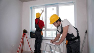 Apartment repair in the room - two men workers measuring the distance from the windowsill to the ceiling and another writing notes about it