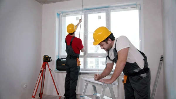 Apartment repair in the room - two men workers measuring the distance from the windowsill to the ceiling and another writing notes about it — 스톡 사진