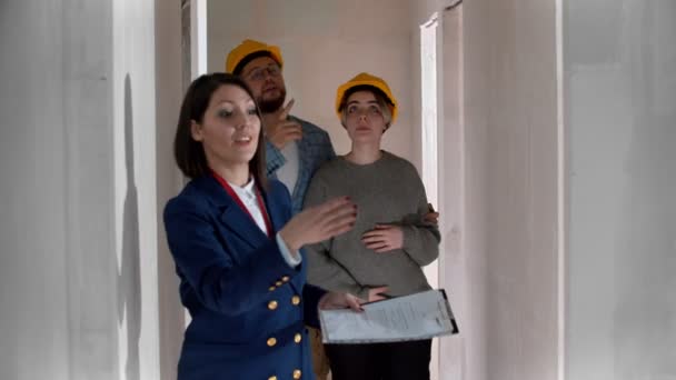 Агент по недвижимости показывает новый проект квартиры молодой супружеской паре в шлемах - оглядываясь вокруг — стоковое видео