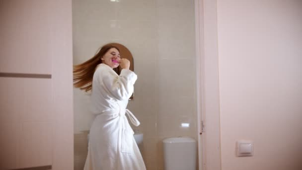 Una joven mujer feliz cepillándose los dientes por la mañana usando un albornoz — Vídeo de stock