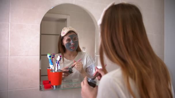 Молода жінка наносить сіру маску на обличчя за допомогою пензлика — стокове відео