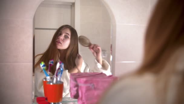 Une jeune femme se brosse les cheveux épais en regardant dans le miroir — Video