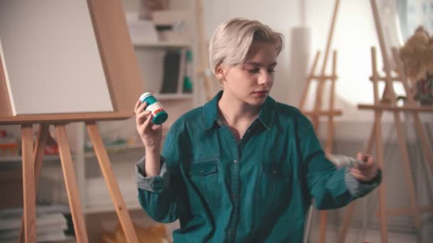 Una joven artista mirando dos tubos diferentes de una pintura azul — Vídeos de Stock