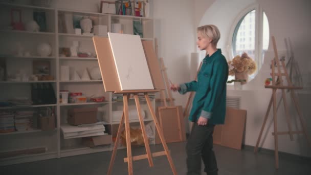 En ung blond kvinna konstnär går till ett staffli och rita skiss på duken — Stockvideo