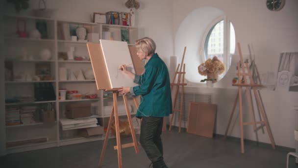 Een jonge blonde vrouw kunstenaar tekenen schets op het doek en luister muziek in de hoofdtelefoon — Stockvideo