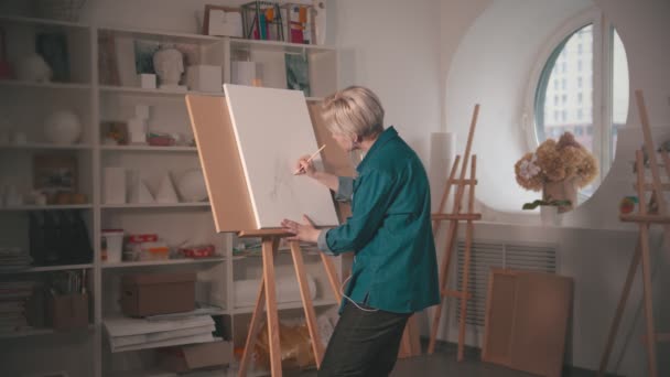 Kısa sarı saçlı genç bir kadın tuvale resim çiziyor ve kulaklıkla müzik dinliyor. — Stok video