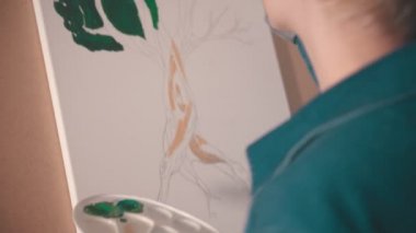 Kısa sarı saçlı genç bir kadın sanat stüdyosunda ağacın üst dallarını yeşile boyuyor.