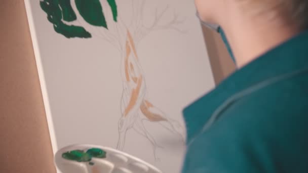 Una joven con pelo corto y rubio pintando ramas superiores de árbol en color verde en el estudio de arte — Vídeo de stock