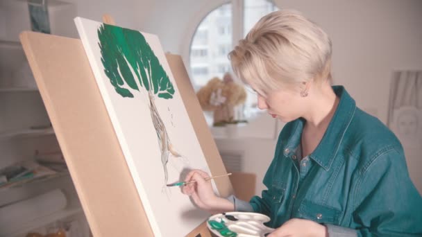 A young blonde woman draws a tree with paints in the art studio — 비디오