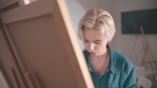 A young blonde woman artist draws a painting in the art studio — Stock Video